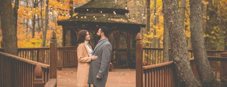 A man and woman standing close in front of the gazebo at Castle in the Country against beautiful fall foliage, gazing at each other after he proposed