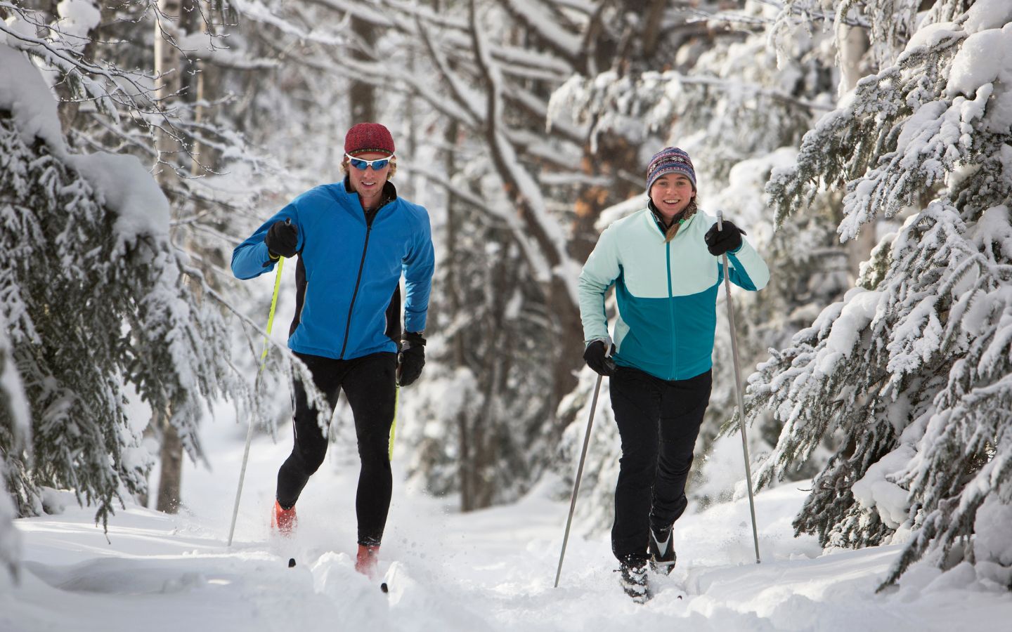 Man and woman smiling and cross country skiing on a trail through a pine forest.
