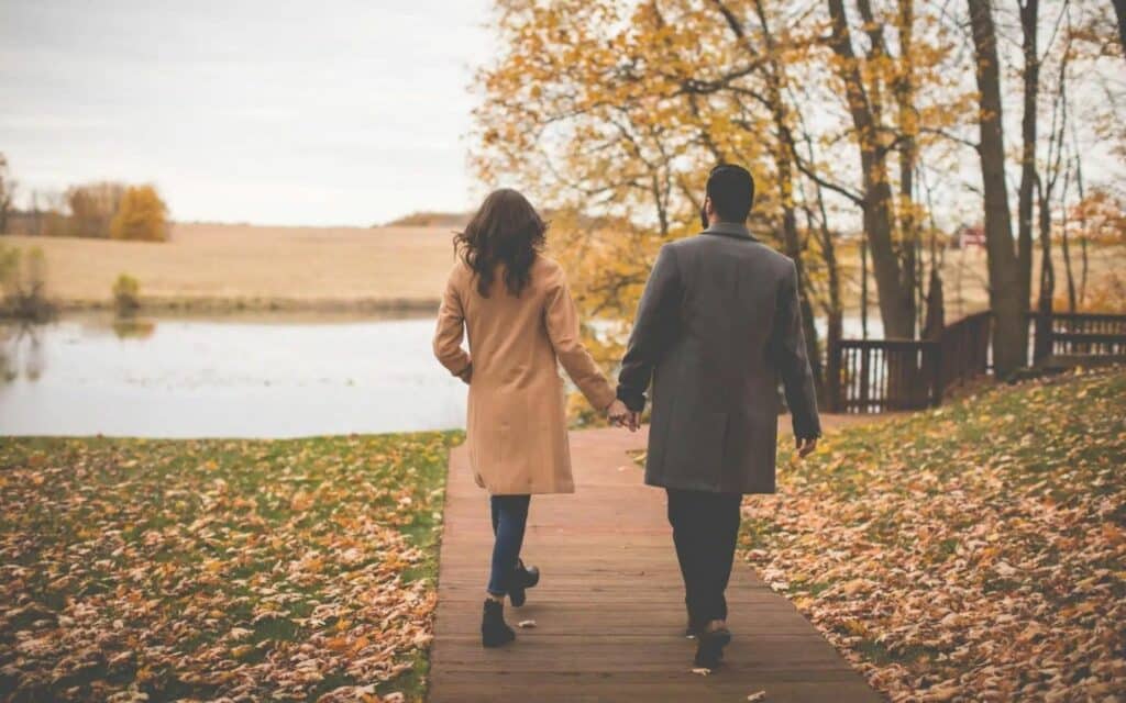 A couple walking hand in hand on a boardwalk toward a pond surrounded by fall foliage