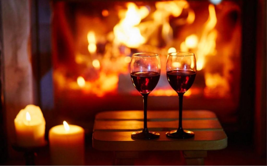 Lit candles and two wine glasses with red wine in front of a glowing fireplace