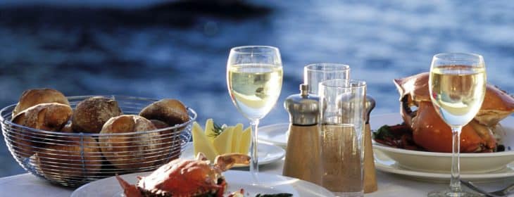 Dining table by the water topped with white linen, seafood and wine
