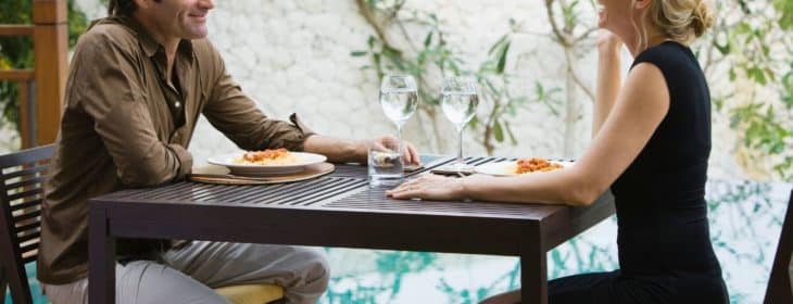Couple dining at a table for two outside with water and greenery in the background