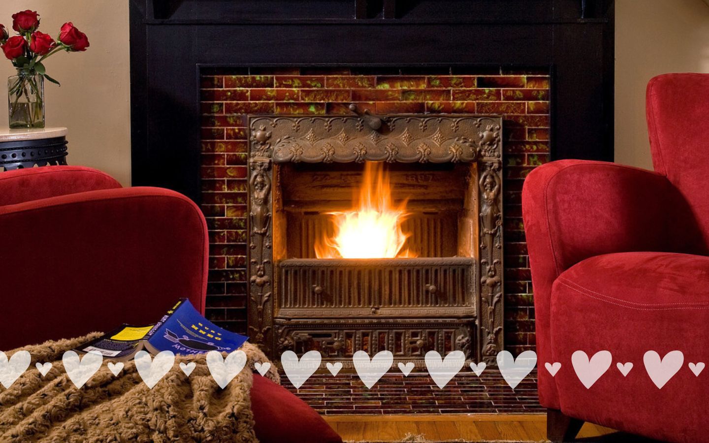 The Crowning Suite at Castle in the Country with a wood-burning fireplace, two red armchairs, and a vase of roses in the corner. Heart stickers across the bottom of the photo.